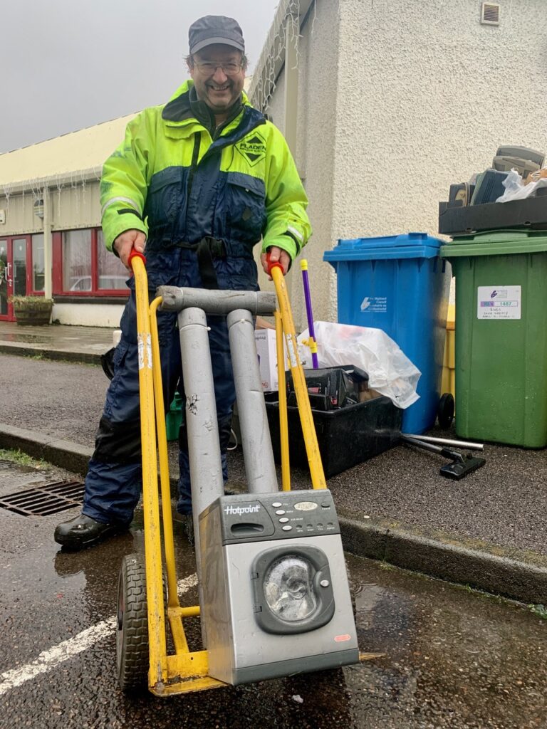 Man in baseball cap and hi vis with mini washing machine on an orange sack troller