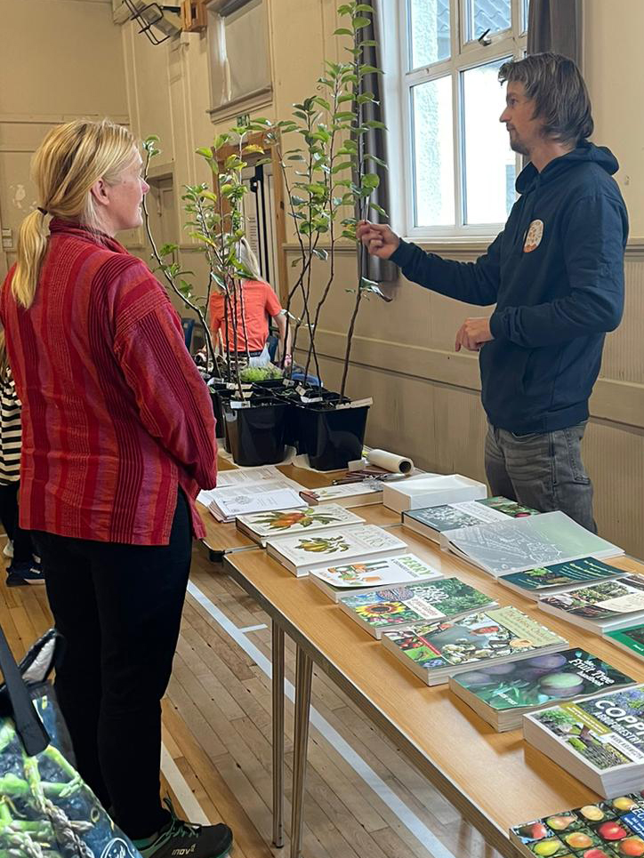 a woman listens to a man showing a small apple tree on a table covered with books